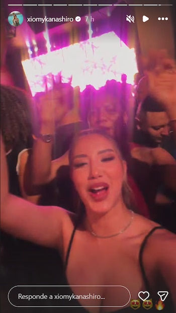 Una mujer sonríe y levanta el brazo mientras graba un video en una fiesta con varias personas bailando al fondo con luces tenues