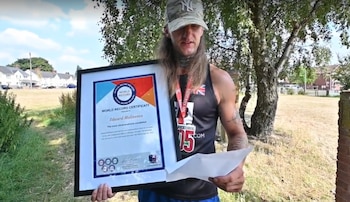 Un hombre con gorra y camiseta sin mangas sostiene un cuadro con un certificado y una hoja de papel al aire libre junto a unos árboles