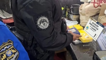 Dos policías con uniformes oscuros inspeccionan paquetes amarillos sobre una mesa oscura. Uno sostiene un dispositivo. Un cartel de "Evidencia N° 1" y "Policía" es visible