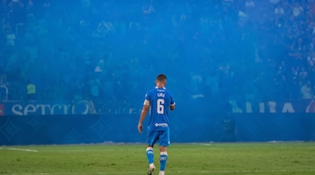 Un jugador de fútbol de espaldas con la camiseta número seis camina en una cancha frente a una tribuna cubierta de humo azul.