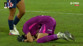 Un guardameta con uniforme morado apoya la cabeza y las manos sobre el césped de una cancha de fútbol