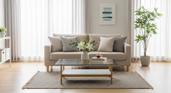 Una sala de estar luminosa y ordenada con un sofá color crema, una mesa de centro de cristal con un jarrón de lirios blancos, libros y una taza de té, y una planta verde alta.