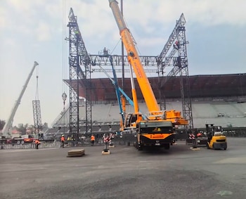 Estadio al aire libre con grúas y trabajadores montando una gran estructura de escenario 360. Se ven asientos vacíos y equipos de construcción