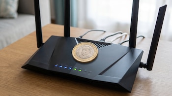 Un router de internet negro con varias antenas y luces indicadoras azules y verdes, sobre el cual descansa una moneda de un euro española, sobre una mesa de madera.