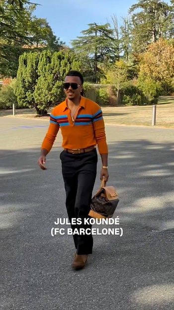 Jules Koundé camina sobre asfalto con gafas de sol, polo naranja con franjas azules, pantalones oscuros y un bolso en la mano