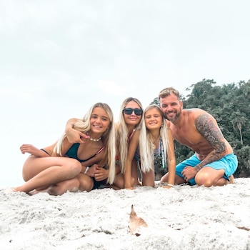 Un hombre, una mujer y dos niñas sonríen arrodillados sobre la arena blanca de una playa con vegetación al fondo