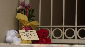 Un ramo de flores envuelto en papel amarillo, dos osos de peluche (uno blanco, uno rojo) y una nota de pésame junto a una reja blanca