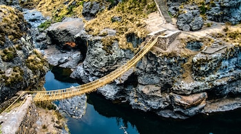 Puente de cuerda que cruza un desfiladero rocoso sobre agua azul, rodeado de vegetación baja y gradas de piedra.