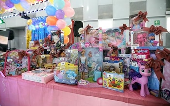 Primer plano de una mesa cubierta con un mantel rosa, repleta de juguetes nuevos en sus cajas, muchos con moños marrones. Globos de colores adornan el fondo