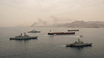 Vista aérea de buques militares y petroleros en el mar. Humo se eleva desde la costa montañosa al fondo bajo un cielo brumoso.