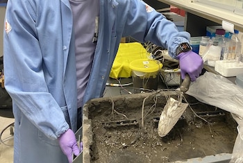 Primer plano de un investigador con guantes morados y bata de laboratorio azul manipulando lodo con una paleta en una bandeja, con cables y equipo de laboratorio