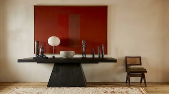 A mesa de console escura tem vasos pretos, luminárias brancas, tigelas cinza e quadros de madeira, contra pinturas vermelhas em paredes bege, com cadeiras e tapetes.