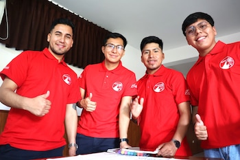 Cuatro hombres jóvenes vestidos con polos rojos iguales posan sonrientes detrás de una mesa con cuadernos y marcadores