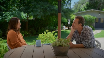 Jason Segel y Cobie Smulders sentados en una mesa de madera al aire libre, ella con cárdigan naranja y él con camisa a cuadros. Una taza y una planta están en la mesa