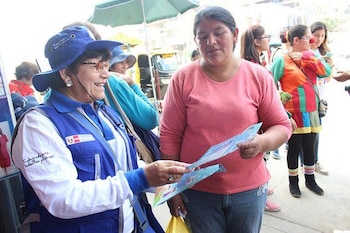 Una mujer sonriente con sombrero y chaleco azul entrega folletos a otra mujer, que viste una blusa rosa y jeans, en una calle con otras personas al fondo