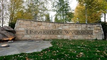Muro de piedra con la inscripción University of Wyoming, rodeado de césped con hojas secas y árboles en el fondo