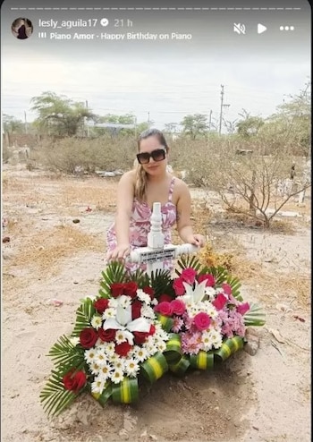 Uma mulher de óculos e vestido floral está ajoelhada em frente a um túmulo com uma pedra branca e duas flores grandes no túmulo. A terra é sujeira