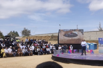 Un hombre con sombrero habla en un escenario circular junto a una pantalla gigante y a un grupo de personas en gradas