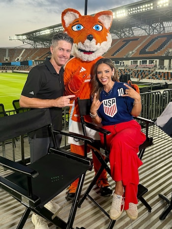 Un hombre, una mujer y una mascota de zorro anaranjada posan sonrientes en un estadio de fútbol con gradas vacías y sillas de director
