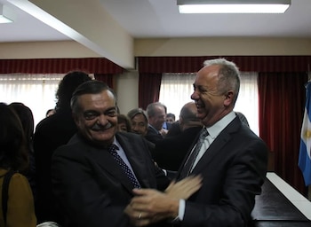 Primer plano de dos hombres vestidos con traje que se sonríen y se tocan los brazos; al fondo, otras personas, cortinas rojas y una bandera argentina