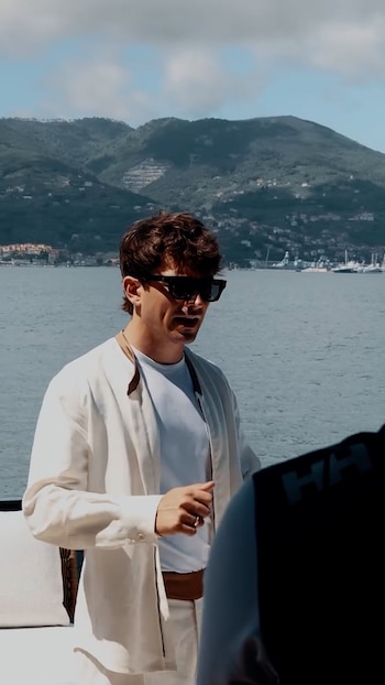 Charles Leclerc, un hombre con gafas de sol y camisa blanca, está de pie en la cubierta de un yate. Detrás de él, hay agua y montañas