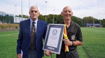 Dos hombres de pie en un campo de fútbol con césped artificial; uno en traje y el otro con uniforme de árbitro, sosteniendo un certificado y una bandera.