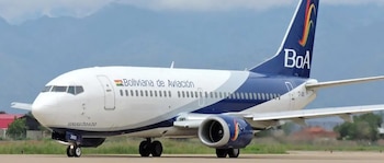 Un avión de Boliviana de Aviación (BoA) blanco y azul está en la pista de un aeropuerto con montañas borrosas y cielo nublado al fondo