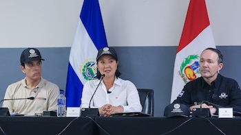 Tres personas sentadas ante micrófonos en una mesa con banderas de El Salvador y Perú de fondo; dos llevan gorras con la inscripción CECOT