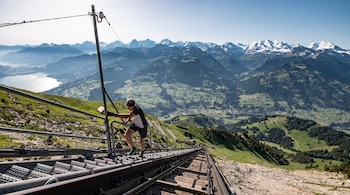 Hombre asciende escaleras metálicas empinadas junto a vía férrea. Detrás, vasto paisaje de montañas, valles verdes, lago y picos nevados bajo cielo azul.