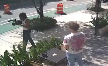 Dos personas en una acera; una de ellas camina portando una cámara, la otra está de espaldas. En el fondo, una calle con conos de tráfico y vegetación