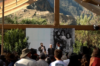 Evento al aire libre bajo una carpa con luces, un escenario con oradores, pantalla con foto en blanco y negro de niños, audiencia, ruinas incas y montañas