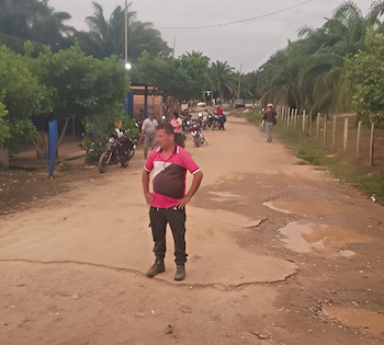 Un hombre de pie en una carretera de tierra con baches y charcos; al fondo, más personas, motocicletas y árboles frondosos bajo un cielo nublado