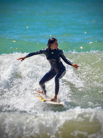 Una mujer con traje de neopreno negro se mantiene de pie sobre una tabla de surf en medio de las olas del mar