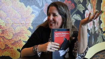 Una mujer sonriente con cabello largo sostiene un libro de tapa roja con el título "Cadáver exquisito" en una mesa frente a un fondo decorado