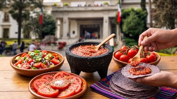 Mesa de madera con un molcajete con salsa, ensalada, rodajas de jitomate, jitomates enteros, tortillas moradas y manos sosteniendo una.