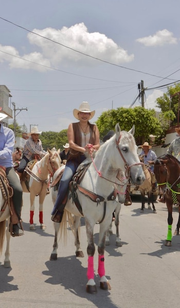 Mujer con sombrero vaquero, chaleco y jeans monta caballo blanco con vendas rosadas en las patas, junto a otras personas a caballo en una calle