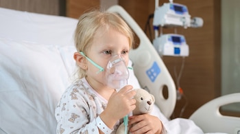 Niña sentada en una cama de hospital con una máscara nebulizadora en el rostro, sosteniendo un peluche junto a equipos médicos.