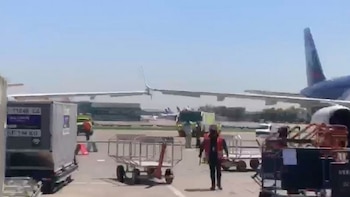 Vista de la plataforma de un aeropuerto con dos aviones, uno con el ala cerca de la cola del otro. Hay personal de tierra y carros de equipaje en primer plano