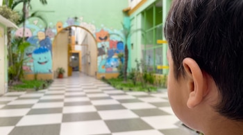 Espalda de la cabeza y oreja de un niño. Fondo desenfocado con pasillo, piso de cuadros blancos y grises, y pared con murales infantiles.