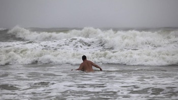 Hombre de espaldas sumergido hasta la cintura en el mar agitado. Grandes olas rompen con espuma blanca bajo un cielo gris y nublado