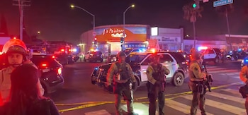 Agentes de policía y vehículos con luces de emergencia encendidas. Cinta amarilla de seguridad en la calle. Un edificio comercial y personas presentes en la escena nocturna