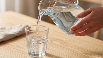 Manos que sirven agua desde una jarra de vidrio hacia un vaso sobre una mesa de madera