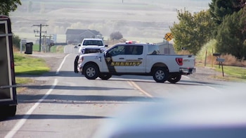 Una camioneta de la policía del alguacil cruza una carretera rural frente a otro vehículo en un paisaje con casas y árboles