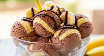 Primer plano de un tazón de vidrio con bolas de helado de chocolate y plátano, adornado con salsa de chocolate, rodajas de plátano y plátanos al fondo.