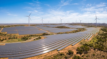 Vista aérea de extensos campos de paneles solares y aerogeneradores en un terreno árido con vegetación baja bajo un cielo despejado.