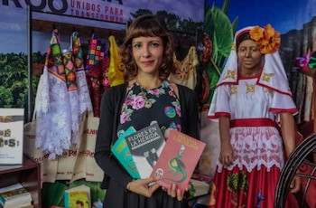 Mujer de cabello castaño con flequillo sostiene tres libros. Detrás, hay un expositor con textiles, un maniquí con ropa tradicional y un cartel