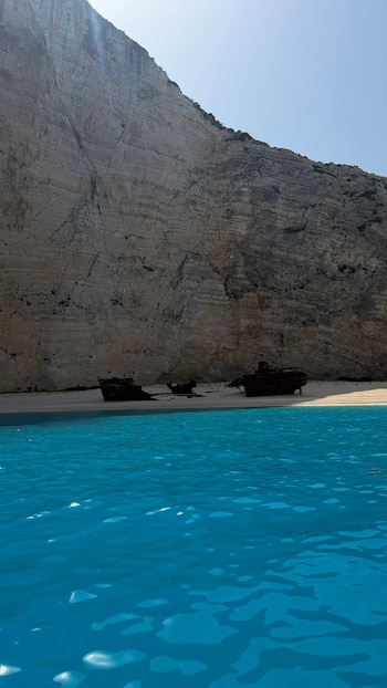 Agua turquesa, playa de arena blanca, dos barcos naufragados oxidados, acantilados grises de roca, cielo azul claro sin nubes