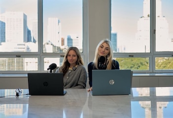 Dos mujeres sentadas ante una mesa con dos computadoras portátiles y dos micrófonos, frente a ventanales con vista a varios edificios