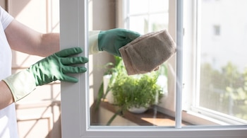 Manos protegidas con guantes verdes limpian con un paño doblado el cristal de una ventana blanca.