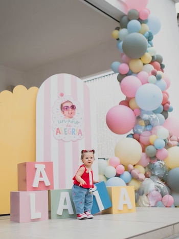 Una niña pequeña está de pie junto a cubos de colores que forman el nombre ALAIA frente a un adorno de globos y carteles